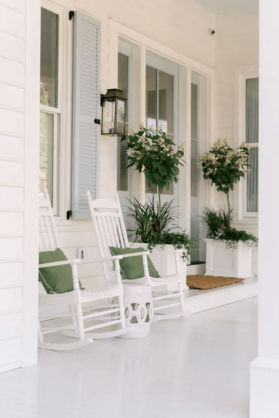 a white rustic summer porch with white furniture and planters with blooms and greenery, green pillows and a side table is very airy