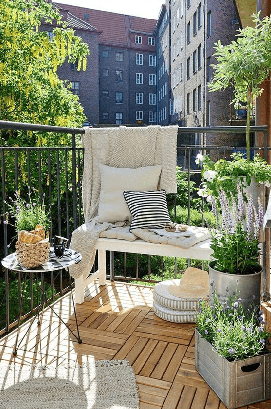 an airy summer balcony with a white bench and printed textiles, a small table, potted blooms and greenery and cushions