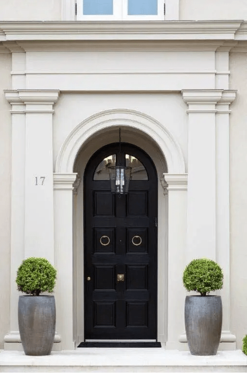 an exquisite entrance with a black arched door and ring handles, tall ceramic pots with topiary trees is a cool and chic space