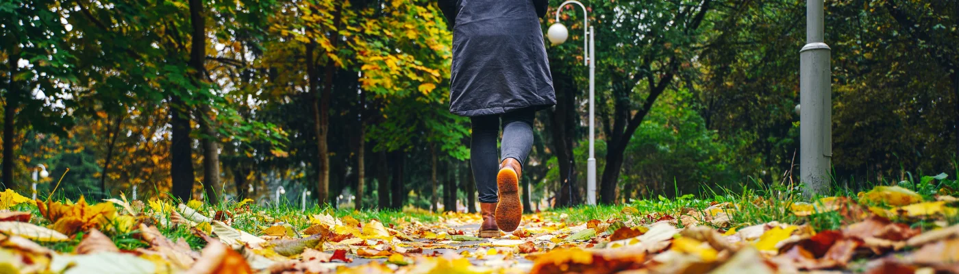 A woman in black jacket walking in a park along the sidewalk strewn with fallen leaves. The concept of turnover seasons. Weather background