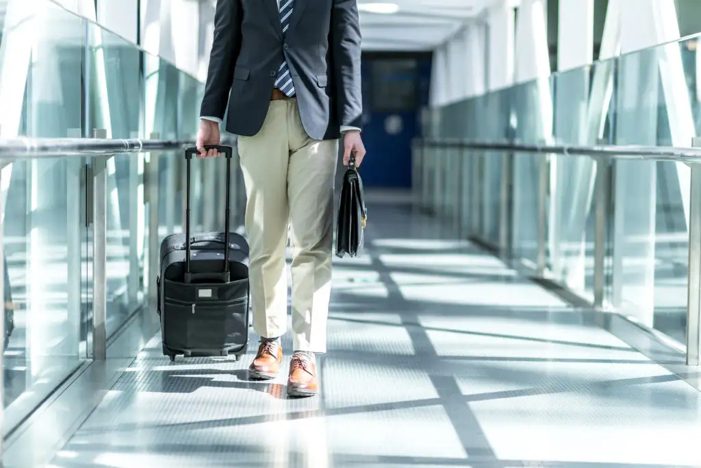 Close up of person walking down glass airport hallway pulling along a carry on suitcase and carrying a briefcase