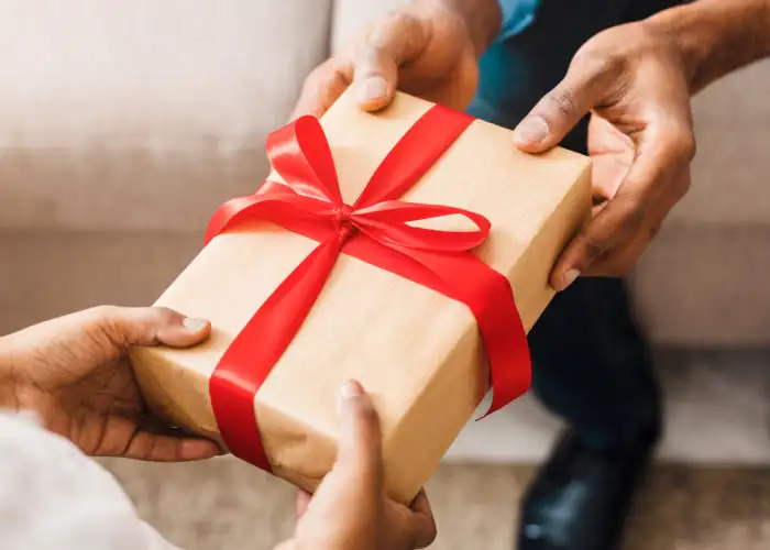 Close up of person handing wrapped gift with a red holiday ribbonto another person