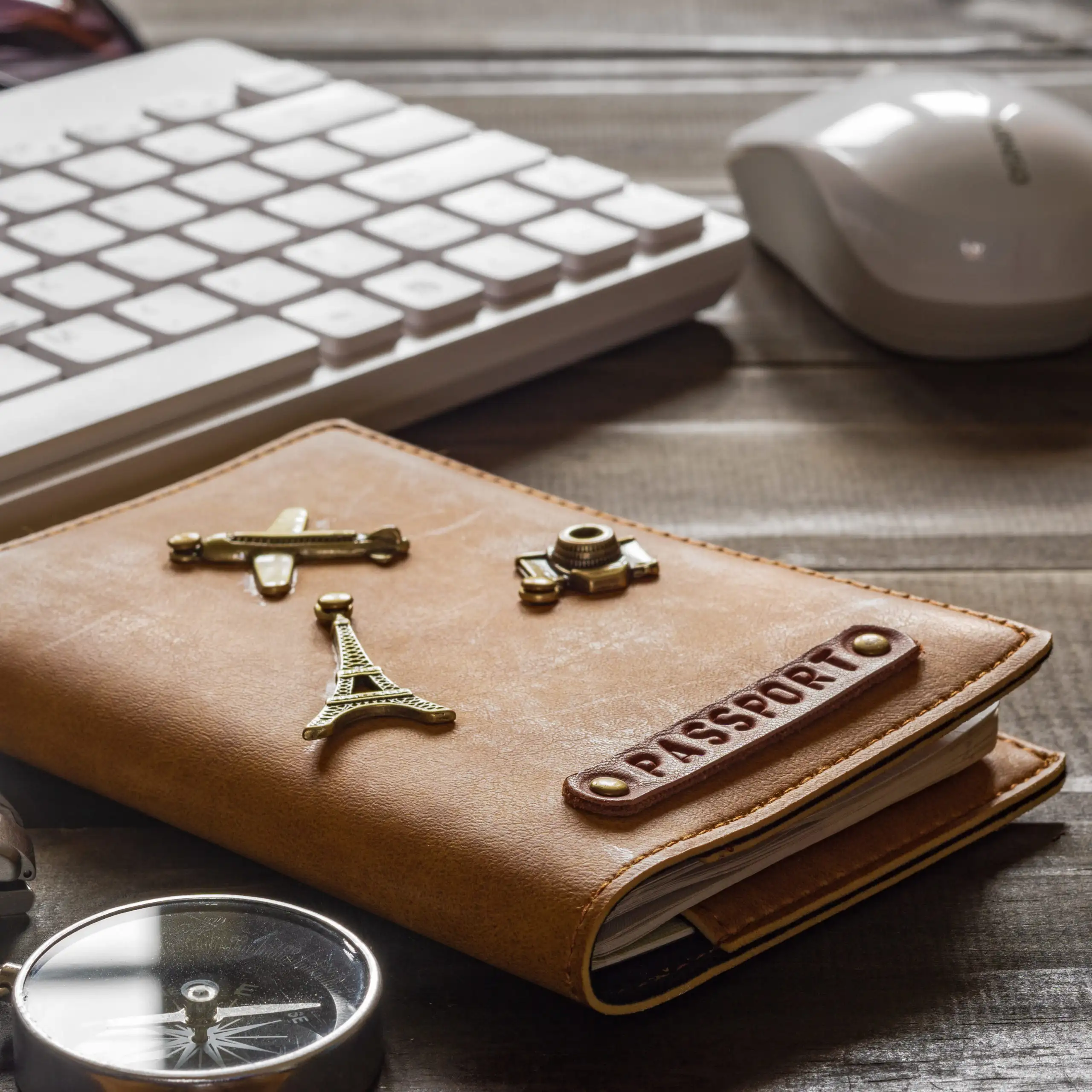 A tan passport cover next to a keyboard, mouse, and compass on a wooden desk