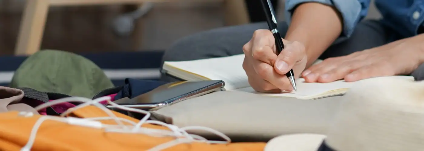 Close up of person writing in notebook surrounded by packed suitcase and various items