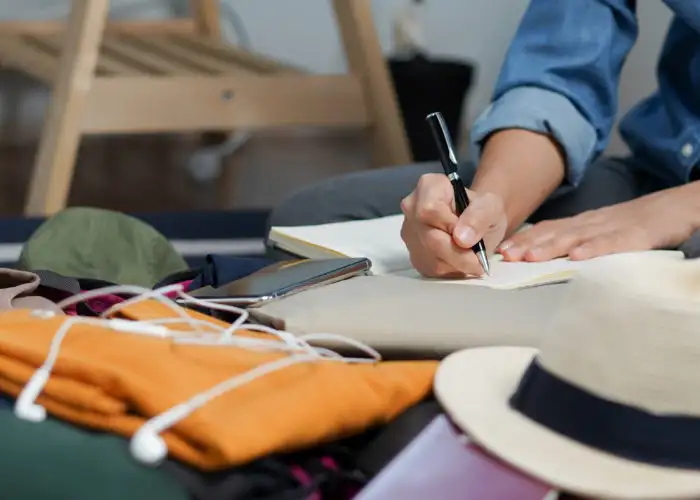 Close up of person writing in notebook surrounded by packed suitcase and various items