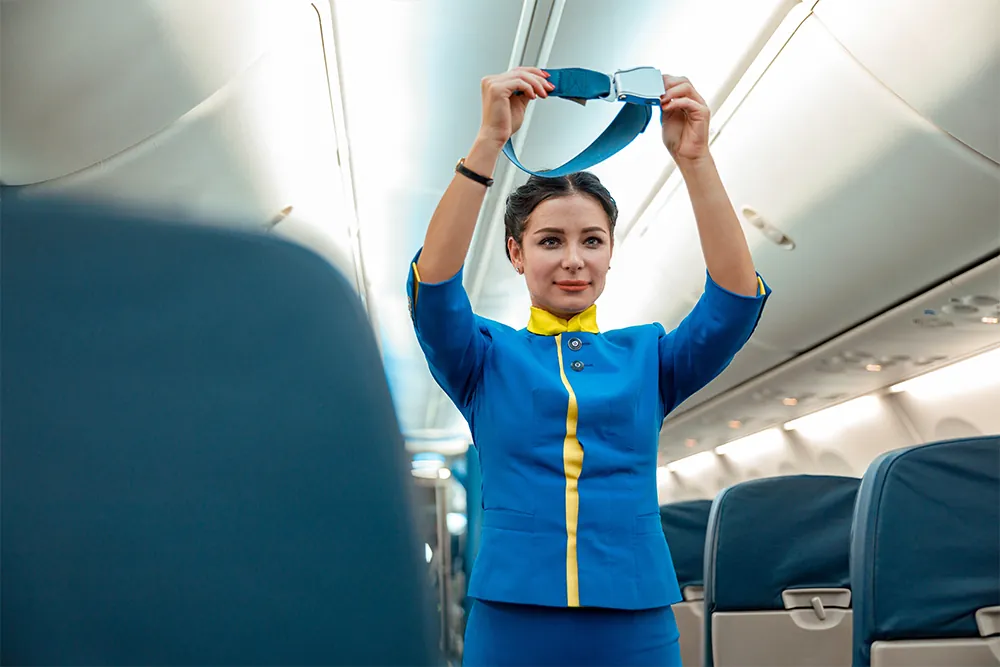 Female flight attendant in air hostess uniform holding seatbelt while standing near passenger seats in airplane