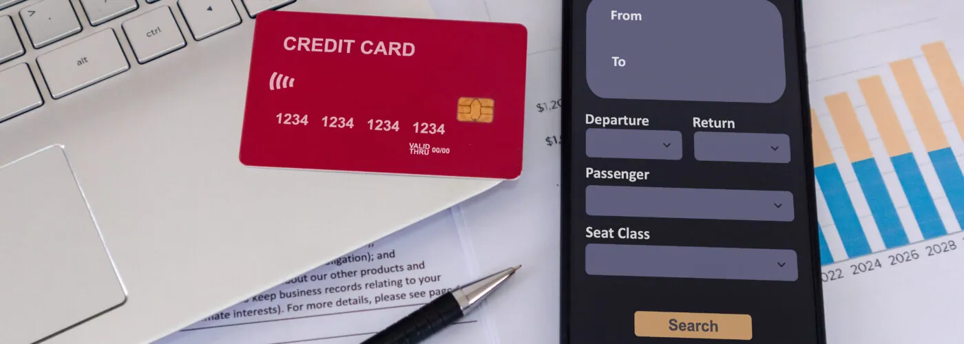 Office desk with credit card, smartphone with online airline ticket booking application on screen, laptop, paperwork, and pen