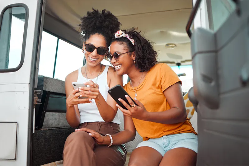 Two friends sitting in the open door of a van using their phones and laughing