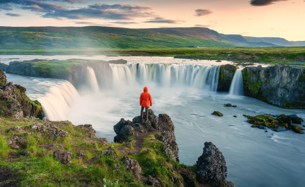 Godafoss waterfall flowing with colorful sunset sky and male tourist standing on cliff in summer at Iceland