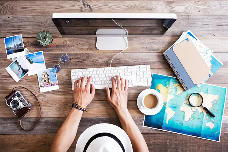 Aerial view of person booking flights surrounded by polaroid photos, maps, a cup of coffee, notebooks, and a camera